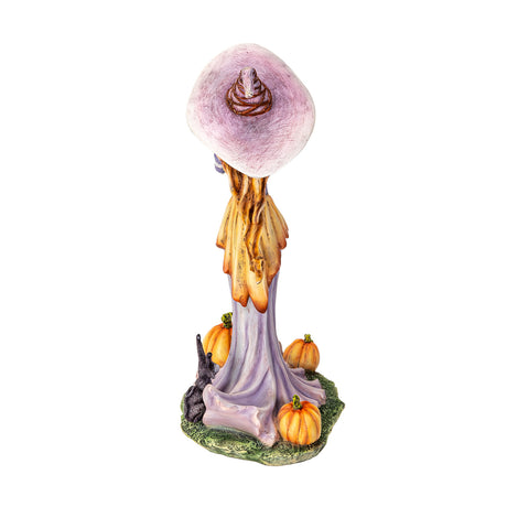 Figurine of a witch in pale purple and orange, holding up a pumpkin with more gourds at her feet and a black rabbit, shown from the back