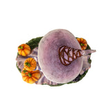 Figurine of a witch in pale purple and orange, holding up a pumpkin with more gourds at her feet and a black rabbit, shown top down