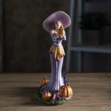 Figurine of a witch in pale purple and orange, holding up a pumpkin with more gourds at her feet and a black rabbit