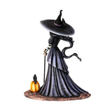 Figurine of a witch in a moody dress and big hat, her hair billowing in an unseen breeze. Next to her is a festive pumpkin, and perched upon it and on her hat are ravens. Shown from the back