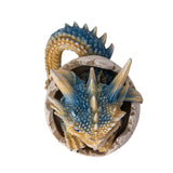 A small fantasy dragon figurine peeking out of a cracked eggshell, shown on a plain white background. The dragon has tan and blue scales, large bright blue eyes, black claws, and small wings. Shown top down