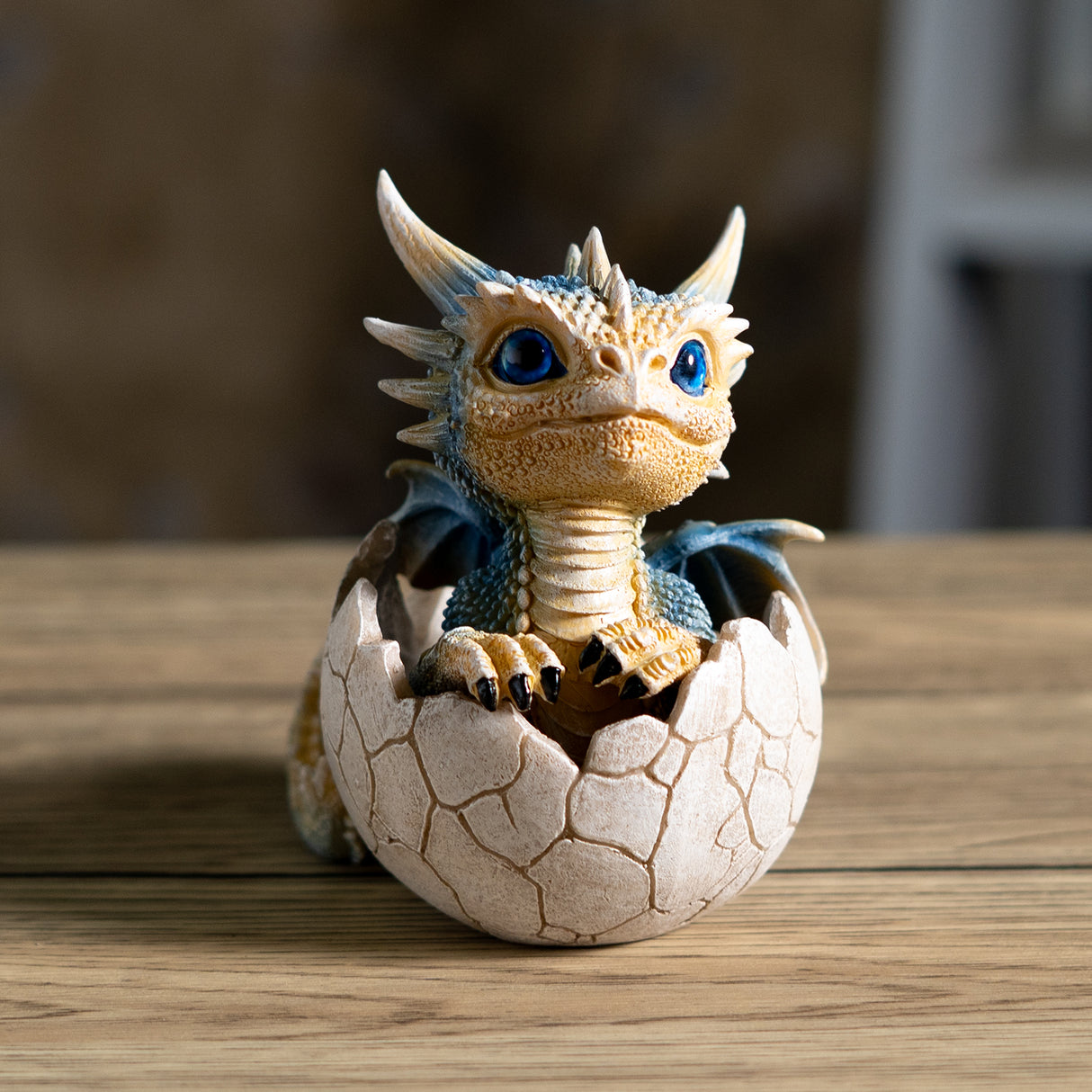 A small fantasy dragon figurine peeking out of a cracked eggshell, shown on a plain white background. The dragon has tan and blue scales, large bright blue eyes, black claws, and small wings.
