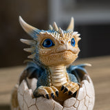 A small fantasy dragon figurine with blue scales and bright blue eyes, emerging from a cracked eggshell on a wooden surface with a blurred background.