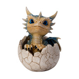 A small fantasy dragon figurine peeking out of a cracked eggshell, shown on a plain white background. The dragon has tan and blue scales, large bright blue eyes, black claws, and small wings.