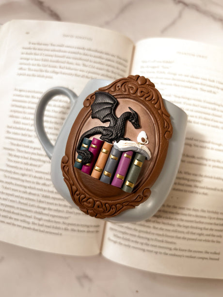 Decorative mug with a dragon and books design shown on an open book background