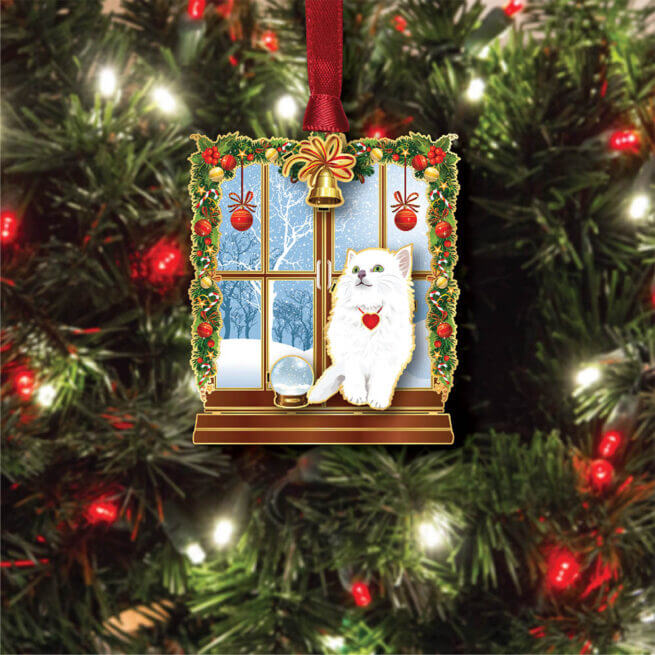 Brass ornament of a white cat sitting in a festive windowsill with snow beyond and a snow globe. shown on a tree