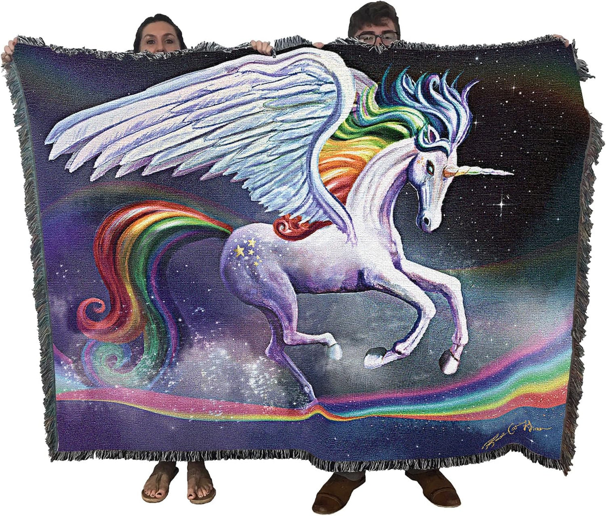 Woven blanket with a unicorn design featuring rainbow colors and a starry background. Unicorn gallops upon a rainbow in the sky. Held by two adults to show large size