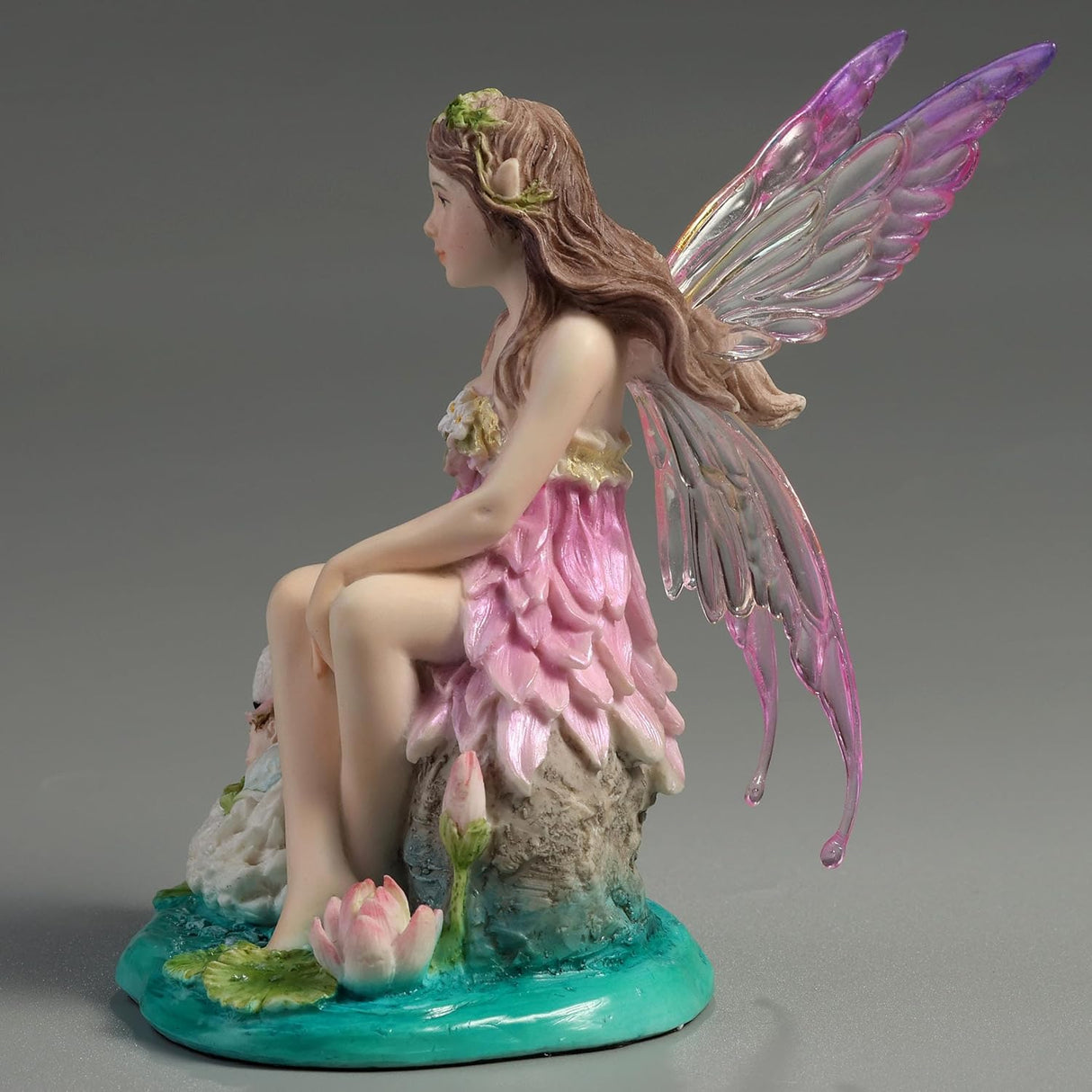 Fairy figurine with pink dress and wings sitting next to a swan with a baby on its back, surrounded by water lilies.