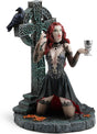Gothic figurine of a vampire woman with red hair holding a goblet in front of a Celtic cross with a raven perched upon it