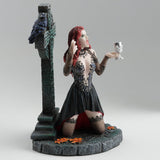 Gothic figurine of a vampire woman with red hair holding a goblet in front of a Celtic cross with a raven perched upon it, shown from the side