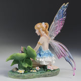 Blond fairy figurine with colorful wings sitting on flowers and grass with a snail on a leaf next to her