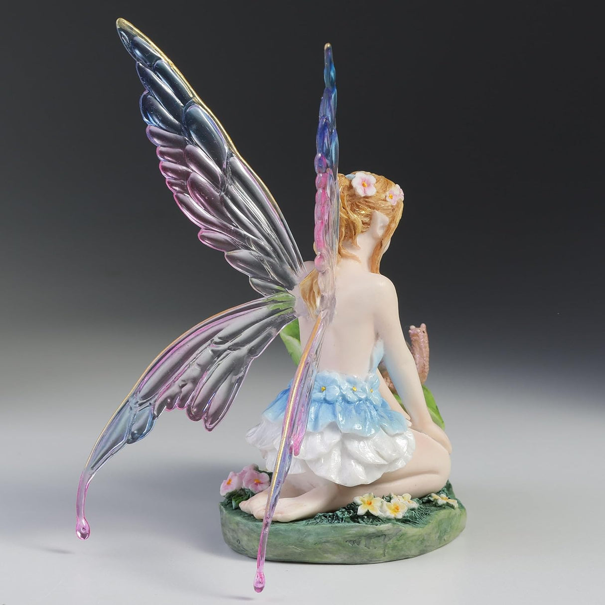 Blond fairy figurine with colorful wings sitting on flowers and grass with a snail on a leaf next to her