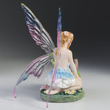 Blond fairy figurine with colorful wings sitting on flowers and grass with a snail on a leaf next to her