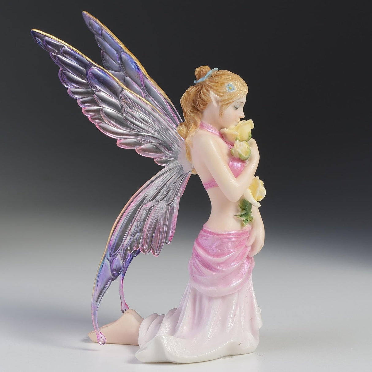 Fairy figurine with colorful wings and a pink dress holding flowers on a gray background