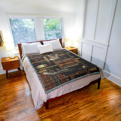 Woven blanket with a design of cats and books with trinkets on a bookshelf. Shown on a bed