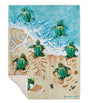 A rectangular throw blanket featuring an illustrated beach scene with green sea turtles swimming in turquoise water and walking on sandy beige shore. The blanket is shown laid flat against a white background, with one corner folded over to reveal a cream underside.