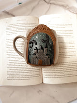 Decorative mug with a castle and dragon design in clay, displayed on an open book