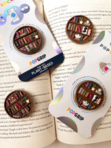 Two miniature bookshelves popsocket phone grips, shown on an open book