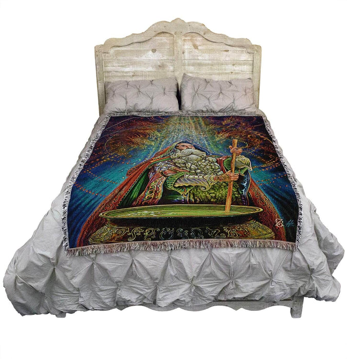 Tapestry blanket with art by Myles Pinkney. Wizard stirring a cauldron with magical dragons in the air above. Shown on a bed