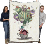 Woven blanket with a colorful design of a dodo in a teacup on hair balloon with dragonflies and butterflies, held up by two adults