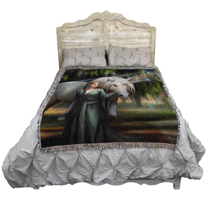 Tapestry blanket with art by Anne Stokes featuring a woman in green with a unicorn standing amidst mossy trees. Shown on a bed
