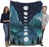 Two people holding a large blanket with a moon phase design over a mountain and forest landscape.