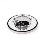 White coaster with black cat with halo sleeping on skulls above word "Saint"