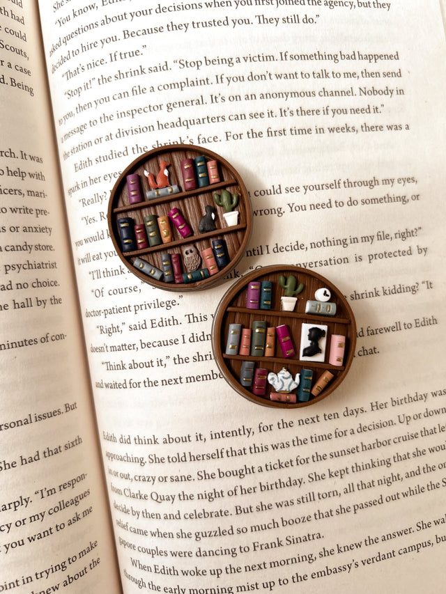 Two miniature bookshelves popsocket phone grips, shown on an open book