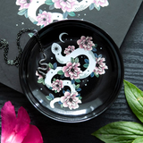 Trinket dish showing white snake with crescent moon, pink flowers on black ceramic background. Shown with a necklace for example