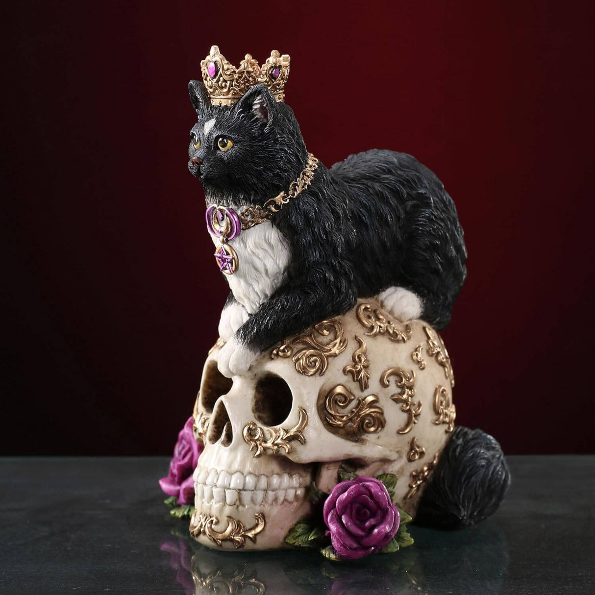 Figurine with a black and white cat in a crown and necklace of gold, sitting on a golden-swirled skull with purple roses