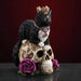 Figurine with a black and white cat in a crown and necklace of gold, sitting on a golden-swirled skull with purple roses