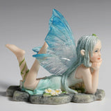 Figurine of fairy in pale blue dress with clear and blue wings and braided hair lying amidst flowers on a stone, feet kicked up