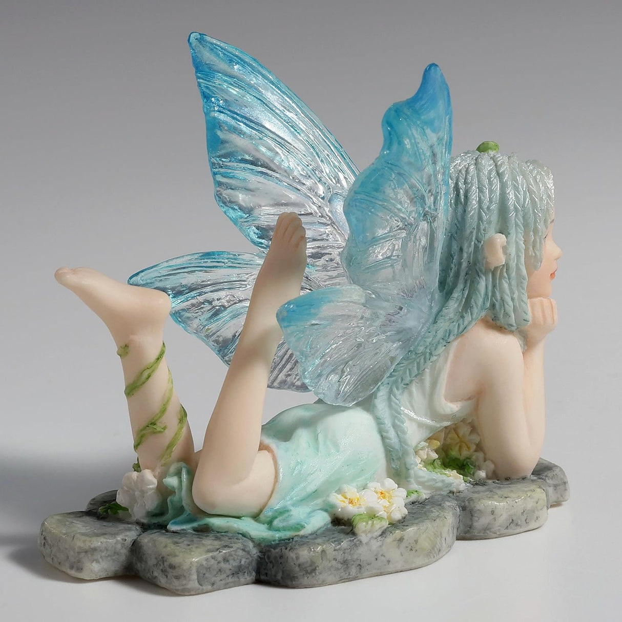 Figurine of fairy in pale blue dress with clear and blue wings and braided hair lying amidst flowers on a stone, feet kicked up
