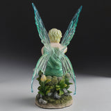 Fairy figurine with green wings, sitting on a base with plants and rocks, shown from the back