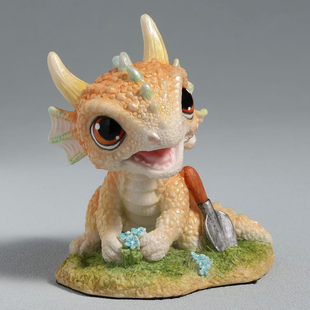 Figurine of a pale gold baby dragon gardening with flowers and a trowel
