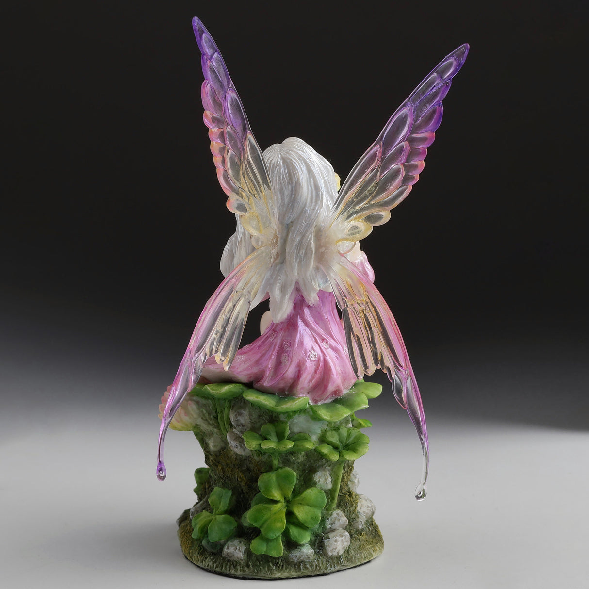 Fairy figurine with purple wings and pink dress sitting amidst clover flowers, shown from the back