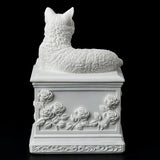 A white cat cremation urn featuring a detailed cat figurine resting on a square pedestal decorated with a central heart-shaped recess and embossed floral rose motifs. Shown from the back