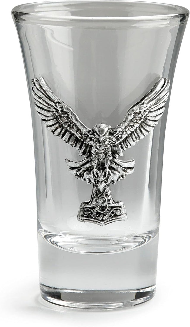 A clear glass shot glass featuring a detailed, raised pewter hawk emblem holding a hammer on the front
