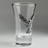 A clear glass shot glass featuring a detailed, raised pewter hawk emblem holding a hammer on the front