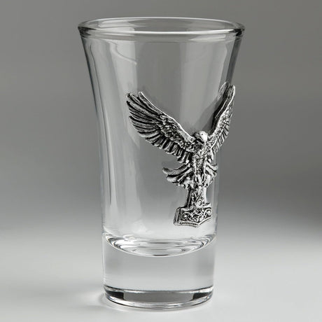 A clear glass shot glass featuring a detailed, raised pewter hawk emblem holding a hammer on the front