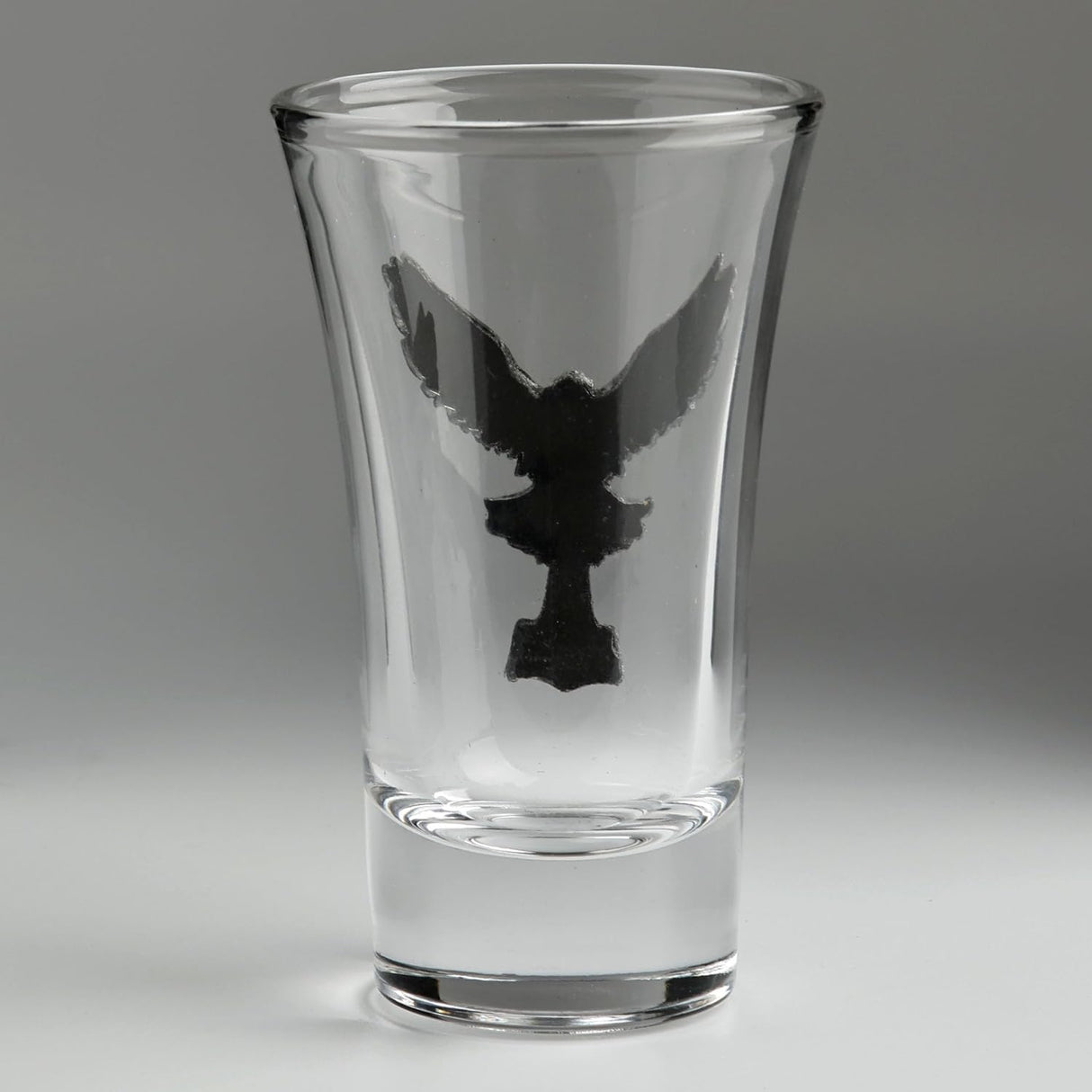 A clear glass shot glass featuring a detailed, raised pewter hawk emblem holding a hammer on the front, seen from the back