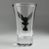 A clear glass shot glass featuring a detailed, raised pewter hawk emblem holding a hammer on the front, seen from the back