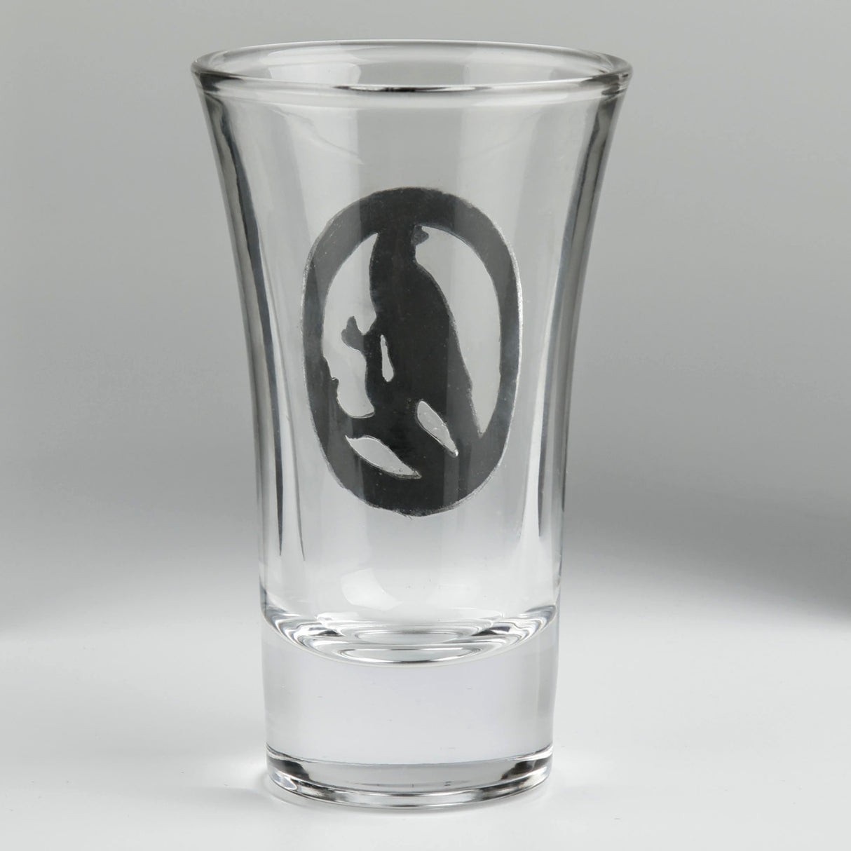 A clear glass shot glass featuring a detailed pewter raven emblem in the center. Shown from the back