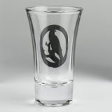 A clear glass shot glass featuring a detailed pewter raven emblem in the center. Shown from the back