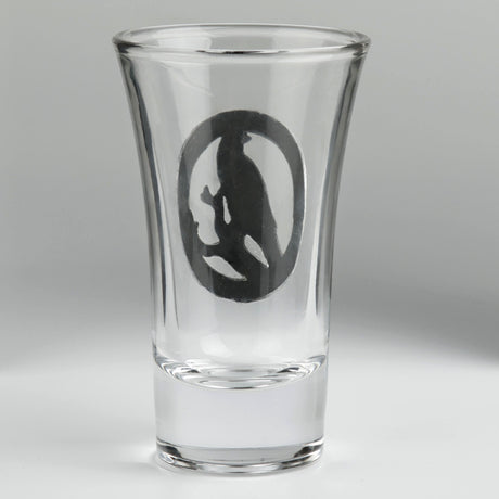 A clear glass shot glass featuring a detailed pewter raven emblem in the center. Shown from the back