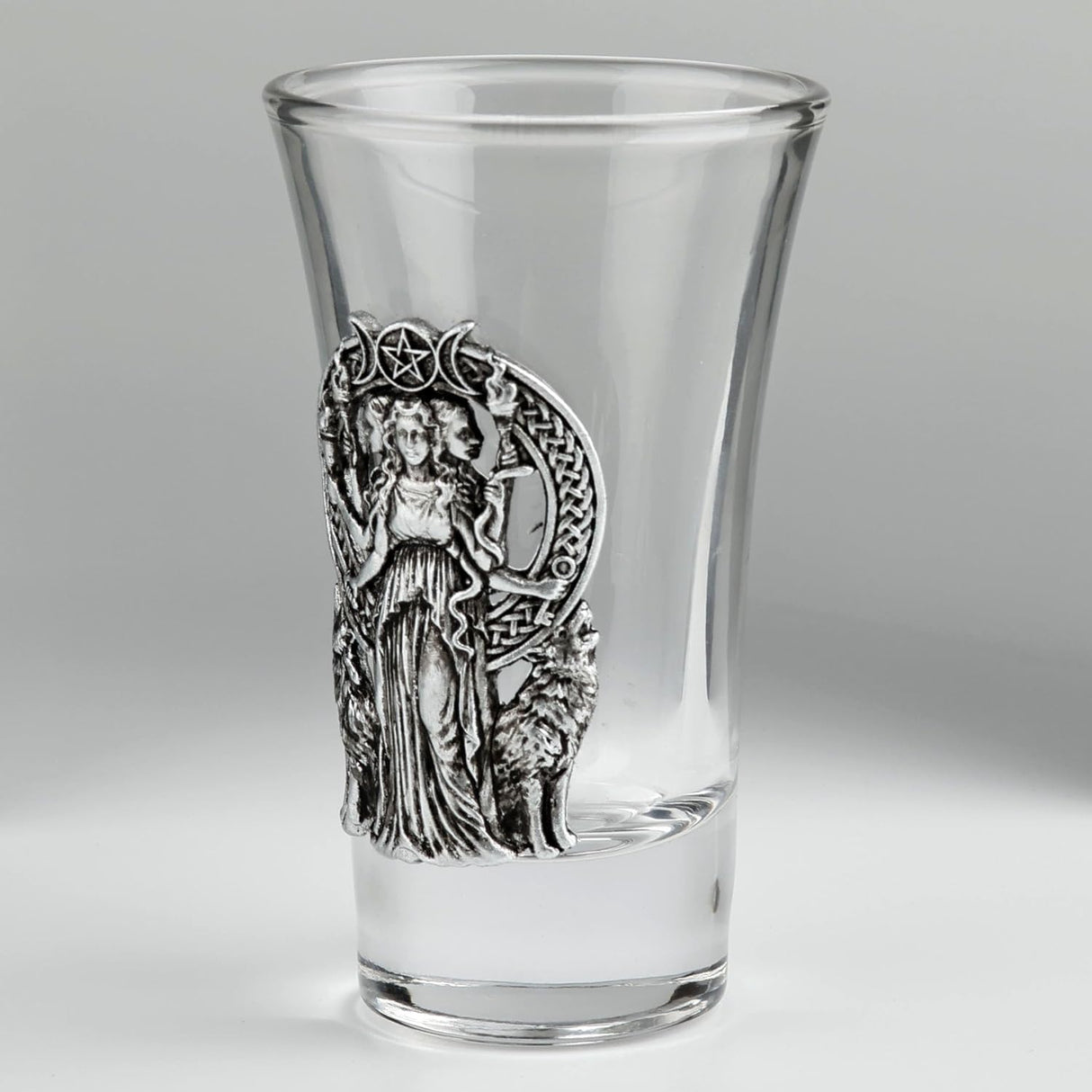 A clear glass shot glass featuring a detailed pewter relief of a triple moon goddess figure, Hecate, flanked by two wolves