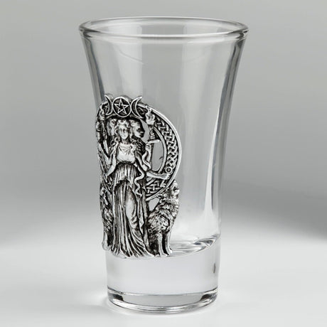 A clear glass shot glass featuring a detailed pewter relief of a triple moon goddess figure, Hecate, flanked by two wolves