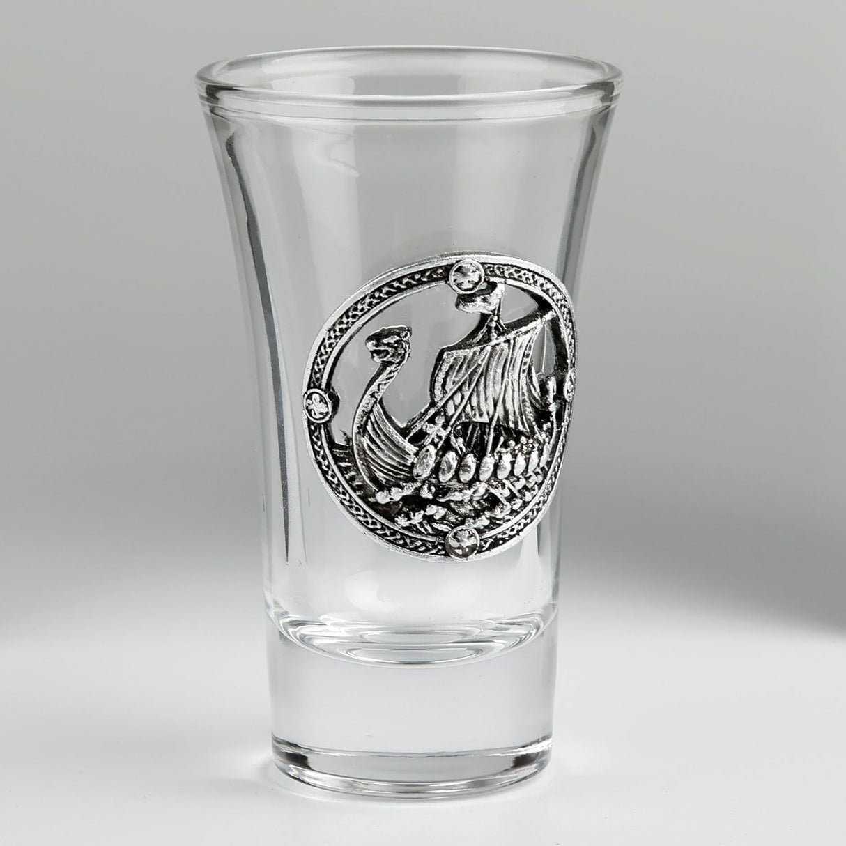 A clear glass shot glass featuring a pewter Viking ship medallion on the front