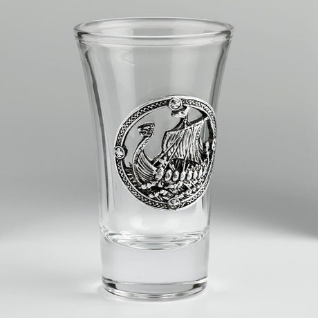 A clear glass shot glass featuring a pewter Viking ship medallion on the front