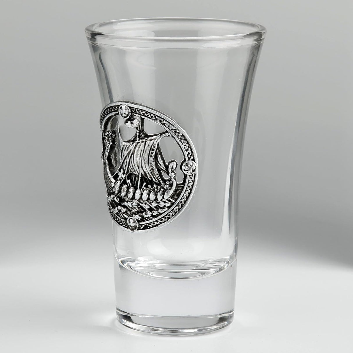 A clear glass shot glass featuring a pewter Viking ship medallion on the front